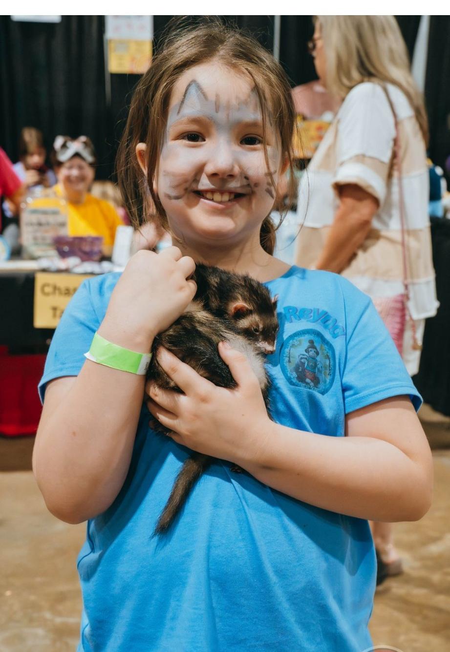 A smiling young girl with face paint gently holding a ferret at an outreach event